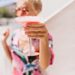 Portrait of elegant lady in bright dress with her hand in focus. Photo of magnificent woman with tanned skin with glass of cocktail on foreground.